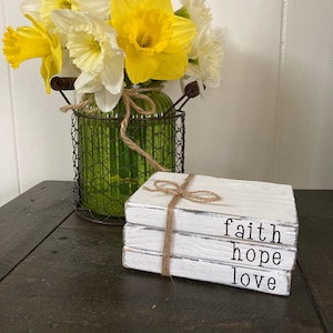 May include: Three white wooden blocks stacked on top of each other with twine tied around them. The blocks are painted white and have the words "faith", "hope", and "love" written on them in black.