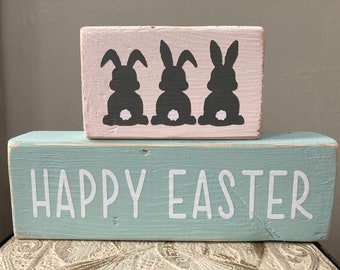 May include: A wooden sign with the text "Happy Easter" in white letters on a light blue background. A smaller wooden sign with three black bunny silhouettes on a pink background sits on top of the larger sign.