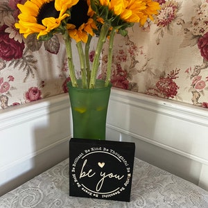 May include: A black wooden sign with a white circle and the words "be you" in gold lettering. The sign is surrounded by a white circle with the words "Be Brilliant, Be Kind, Be Thoughtful, Be Worthy, Be Beautiful, Be Brave, Be Bold" in white lettering.