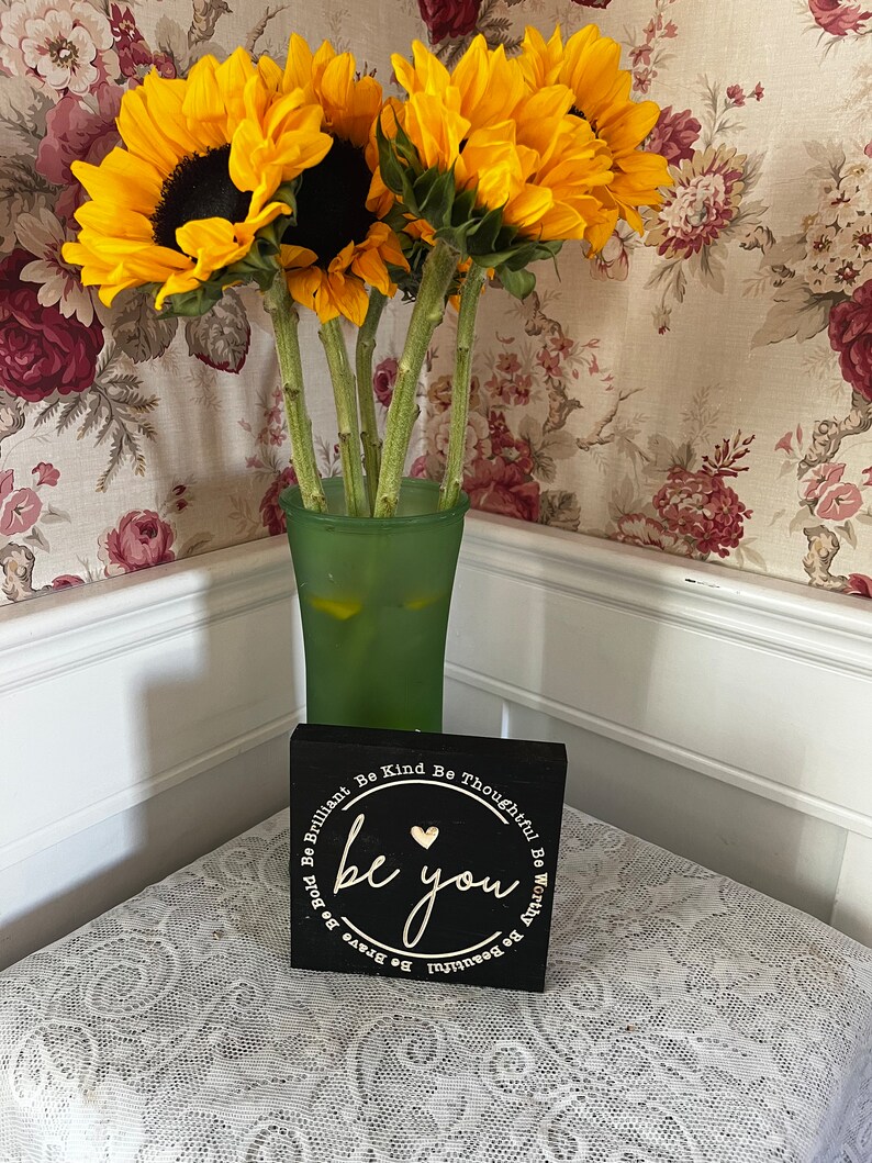 May include: A black wooden sign with a white circle and the words "be you" in gold lettering. The sign is surrounded by a white circle with the words "Be Brilliant, Be Kind, Be Thoughtful, Be Worthy, Be Beautiful, Be Brave, Be Bold" in white lettering.