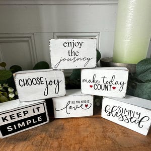 May include: A collection of white wooden blocks with black lettering. The blocks display inspirational phrases like "Enjoy the Journey," "Choose Joy," and "Keep it Simple." Decorative items for home decor.