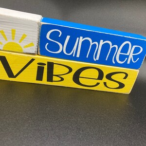 Pode incluir: Tr&ecirc;s blocos de madeira empilhados um em cima do outro. O bloco superior &eacute; branco com um gr&aacute;fico de sol amarelo. O bloco do meio &eacute; azul com a palavra "Summer" escrita em branco. O bloco inferior &eacute; amarelo com a palavra "Vibes" escrita em preto.