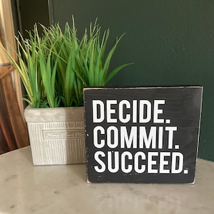 May include: Black wooden sign with white text that reads "Decide. Commit. Succeed." The sign is sitting on a white marble surface next to a potted green plant.