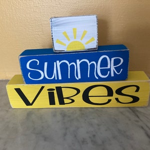 Pode incluir: Tr&ecirc;s blocos de madeira empilhados um em cima do outro. O bloco superior &eacute; branco com um sol amarelo. O bloco do meio &eacute; azul com a palavra "Summer" em tinta branca. O bloco inferior &eacute; amarelo com a palavra "Vibes" em tinta preta.