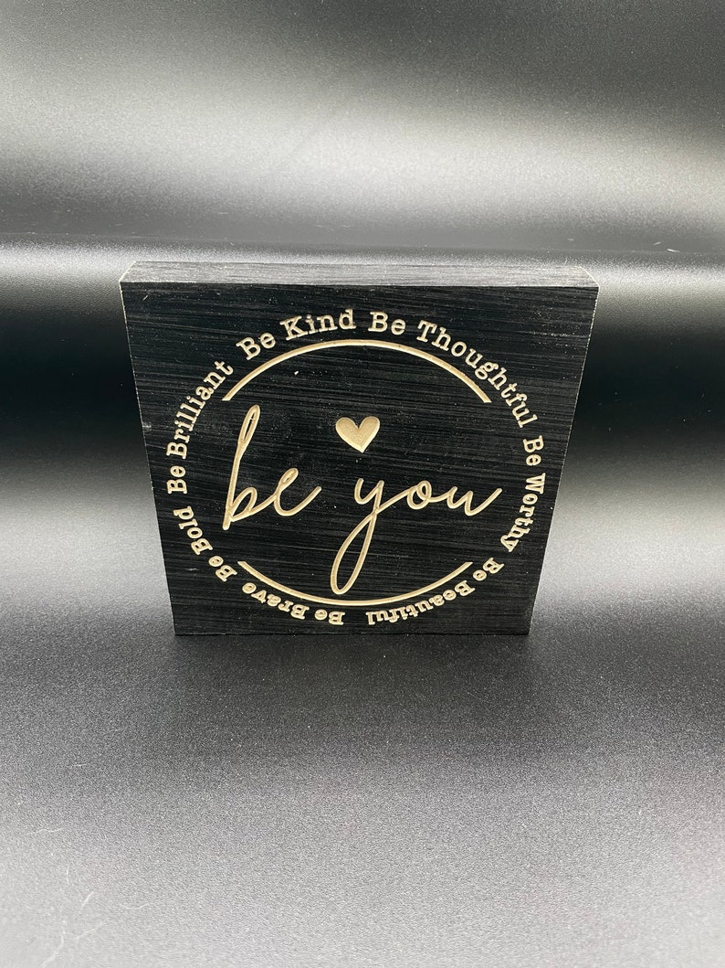 May include: Black wooden block with a gold engraved circle that says "be you" with a heart in the centre. The circle is surrounded by the words "Be Kind Be Thoughtful Be Worthy Be Beautiful Be Brave Be Bold Be Brilliant".