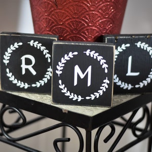 May include: Three black wooden blocks with white letters R, M, and L, each encircled by a white laurel wreath design. The blocks are displayed on a decorative black metal stand, with a red textured vase in the background.
