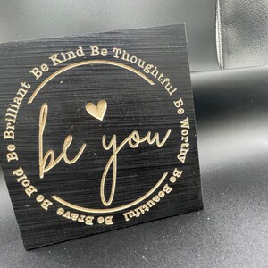 May include: Black wooden sign with gold lettering that reads "Be Kind Be Thoughtful Be Worthy Be Beautiful Be Brave Be Bold Be Brilliant be you". A small heart is in the centre of the circle.