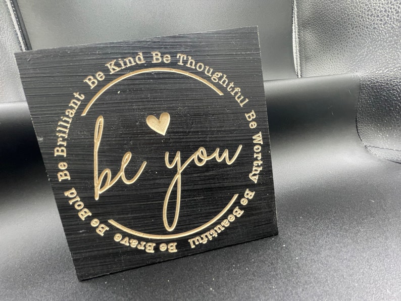 May include: Black wooden sign with gold lettering that reads "Be Kind Be Thoughtful Be Worthy Be Beautiful Be Brave Be Bold Be Brilliant be you". A small heart is in the centre of the circle.