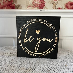 May include: Black wooden sign with gold lettering that reads "Be Bold Be Brilliant Be Kind Be Thoughtful Be Worthy Be Beautiful be you Be Brave".