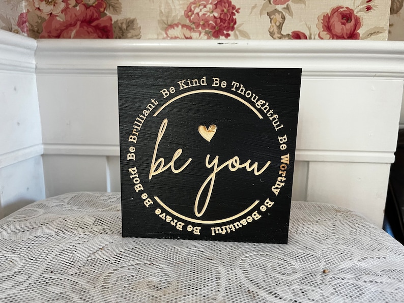 May include: Black wooden sign with gold lettering that reads "Be Bold Be Brilliant Be Kind Be Thoughtful Be Worthy Be Beautiful be you Be Brave".