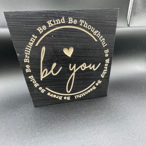 May include: Black wooden sign with a gold circle and the words "Be Brave Be Bold Be Brilliant Be Kind Be Thoughtful Be Worthy Be Beautiful" surrounding the words "be you" in a gold script font.