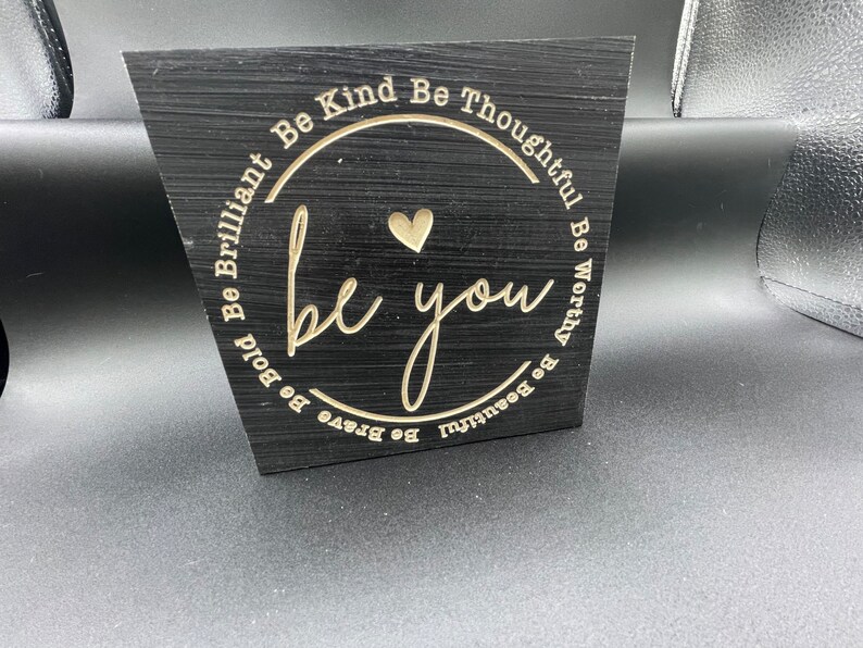 May include: Black wooden sign with a gold circle and the words "Be Brave Be Bold Be Brilliant Be Kind Be Thoughtful Be Worthy Be Beautiful" surrounding the words "be you" in a gold script font.