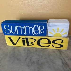 Pode incluir: Blocos de madeira empilhados com as palavras "Summer Vibes" pintadas em letras brancas e pretas sobre um fundo azul e amarelo. Um pequeno bloco branco com um gr&aacute;fico de sol amarelo est&aacute; no meio.