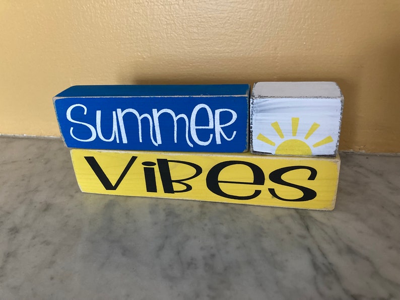 Pode incluir: Blocos de madeira empilhados com as palavras "Summer Vibes" pintadas em letras brancas e pretas sobre um fundo azul e amarelo. Um pequeno bloco branco com um gr&aacute;fico de sol amarelo est&aacute; no meio.
