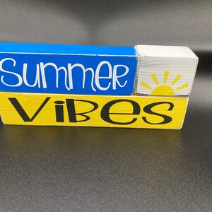 Pode incluir: Um sinal de bloco de madeira com as palavras "Summer Vibes" em tinta preta e branca. O bloco superior &eacute; azul, o bloco do meio &eacute; branco com um sol amarelo e o bloco inferior &eacute; amarelo.
