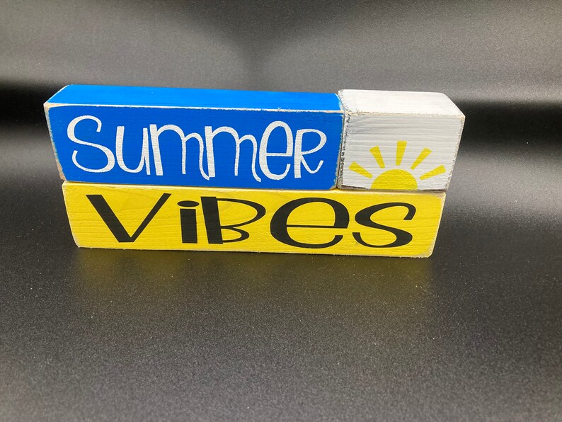 Pode incluir: Um sinal de bloco de madeira com as palavras "Summer Vibes" em tinta preta e branca. O bloco superior &eacute; azul, o bloco do meio &eacute; branco com um sol amarelo e o bloco inferior &eacute; amarelo.