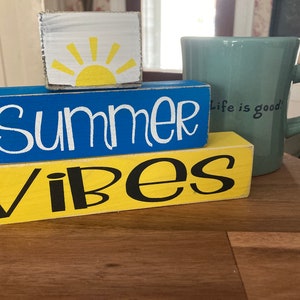 Pode incluir: Blocos de madeira empilhados com as palavras "Summer Vibes" em letras brancas e pretas. O bloco superior tem um sol amarelo com raios brancos sobre um fundo branco. Uma caneca turquesa com o texto "Life is good" est&aacute; no fundo.