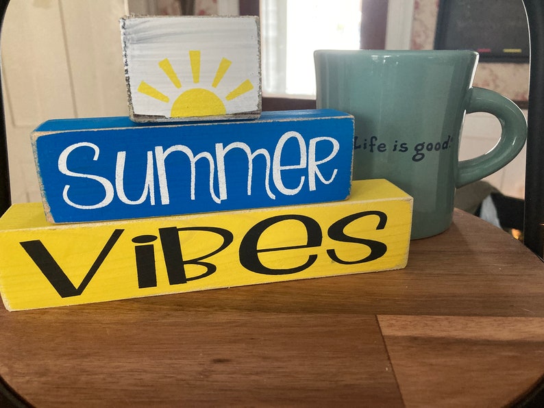 Pode incluir: Blocos de madeira empilhados com as palavras "Summer Vibes" em letras brancas e pretas. O bloco superior tem um sol amarelo com raios brancos sobre um fundo branco. Uma caneca turquesa com o texto "Life is good" est&aacute; no fundo.