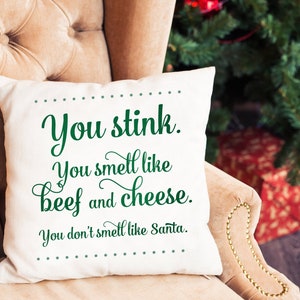 May include: A square, off-white decorative pillow with the text "You stink. You smell like beef and cheese. You don't smell like Santa" in green script. The pillow is on a beige armchair, with a Christmas tree and presents in the background.