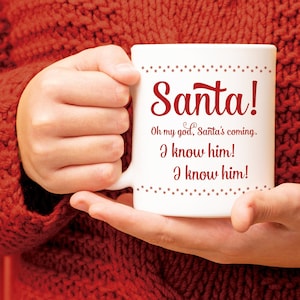 May include: White ceramic mug with the text "Santa!" in red script. Below, the mug reads "Oh my god, Santa's coming. I know him! I know him!" The mug is held by a person wearing a red sweater.
