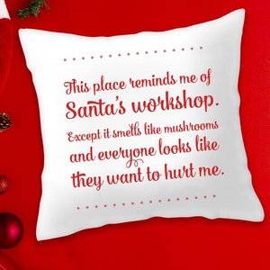 May include: White square cushion with red text: "This place reminds me of Santa's workshop. Except it smells like mushrooms and everyone looks like they want to hurt me." Christmas decorations on a red background.