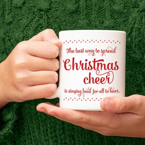 May include: White ceramic mug with the text "The best way to spread Christmas cheer is singing loud for all to hear" in red script. The mug is held by a person wearing a green sweater.