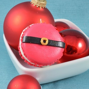 May include: A pink macaron with a black belt and gold buckle, surrounded by red Christmas ornaments in a white bowl.