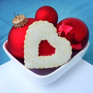 May include: A white heart-shaped cookie with a red jelly center, surrounded by three red Christmas ornaments in a white bowl.