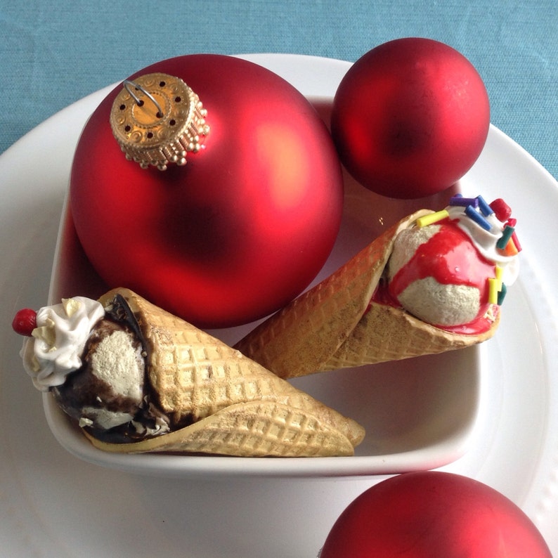 May include: Two red Christmas ornaments and two waffle cones filled with ice cream. One cone has chocolate ice cream with whipped cream and a cherry on top. The other cone has vanilla ice cream with red sauce and sprinkles.