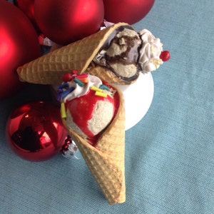 May include: Two ice cream cones with different toppings. One cone has chocolate ice cream with chocolate sauce and whipped cream. The other cone has strawberry ice cream with sprinkles and a red cherry.