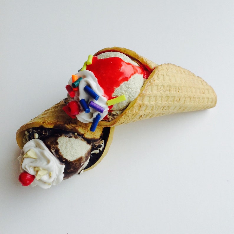 May include: Two scoops of ice cream in a waffle cone. One scoop is vanilla with red sauce and sprinkles, the other is chocolate with whipped cream and a cherry.