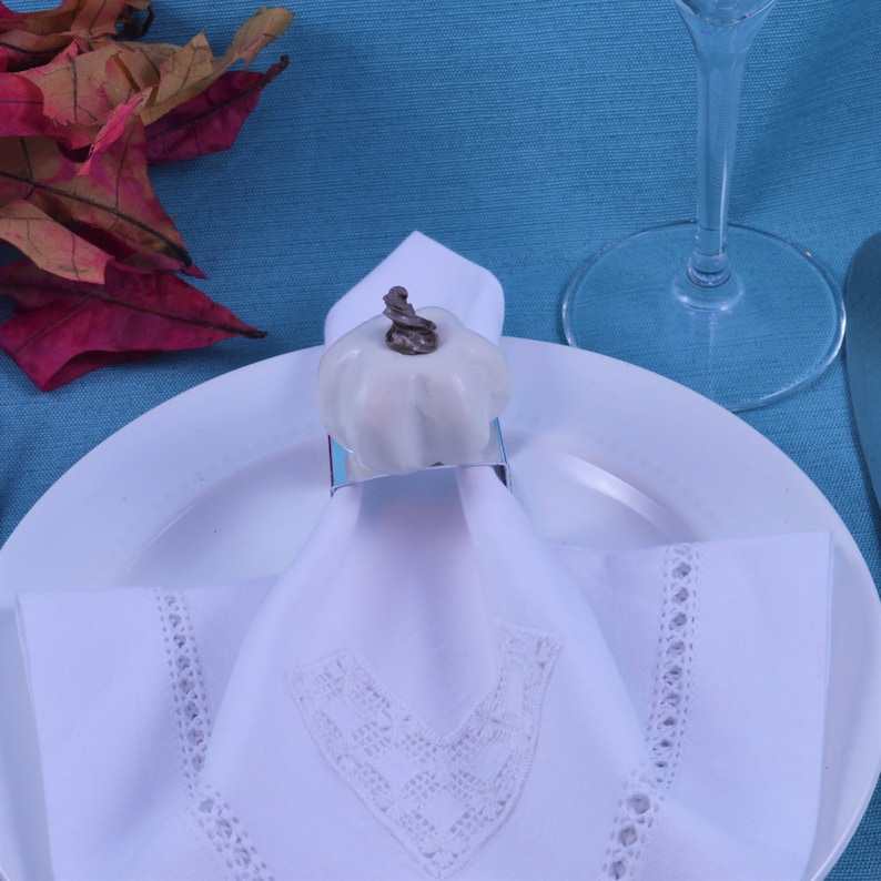 May include: White napkin ring with a white ceramic pumpkin design. The napkin ring is on a white napkin with a lace trim. The napkin is on a white plate.