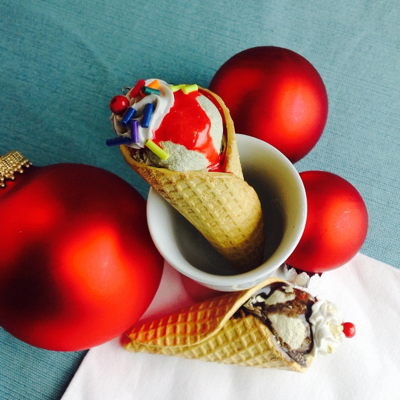 May include: Two ice cream cones with red and white frosting, sprinkles, and a waffle cone. The cones are sitting on a white surface with red ornaments.