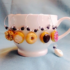 May include: Set of six donut-shaped wine glass charms with black beads. The charms are made of polymer clay and feature different donut toppings, including sprinkles, chocolate, and nuts.