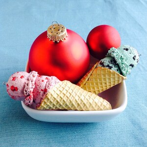 May include: Two ice cream cones with red and green ice cream, topped with a red and a green ornament. The ornaments are hanging from a string.