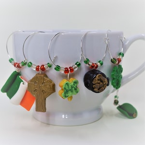 May include: Set of four wine glass charms featuring Irish-themed designs. The charms are made of polymer clay and feature a shamrock, a pot of gold, a Celtic cross, and an Irish flag. The charms are attached to silver-coloured wire rings with red and green beads.