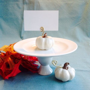 Peut inclure: Citrouilles en céramique blanche avec des porte-cartes en fil d'or sur un support de gâteau blanc. Les citrouilles sont sur un fond bleu.