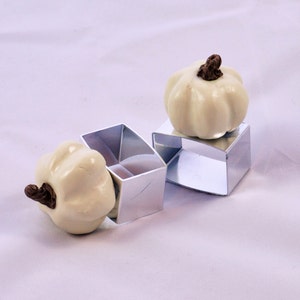 May include: Two white ceramic pumpkins with brown stems, sitting in silver metal napkin rings.