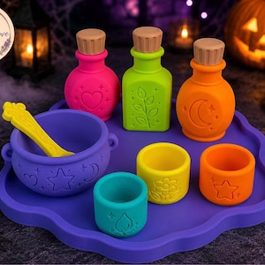 May include: A purple tray holding a purple cauldron with a yellow spoon, three colorful bottles with tan stoppers, and three small cups. The items feature embossed symbols including hearts, stars, moons, leaves, flowers, and flames in bright colors.