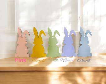 May include: Five wooden bunny shapes in different colors: pink, yellow, green, purple, and blue. The bunnies are arranged in a row on a wooden surface. The colors are labeled below each bunny: Pink, Marigold, Sage, Wisteria, Colonial.