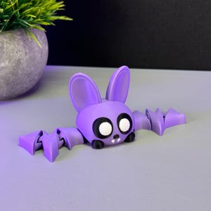 May include: A purple bat figurine with large white eyes and black accents. The bat has articulated wings and a cute, cartoonish design. The figurine is displayed on a light purple surface, with a potted plant in the background.