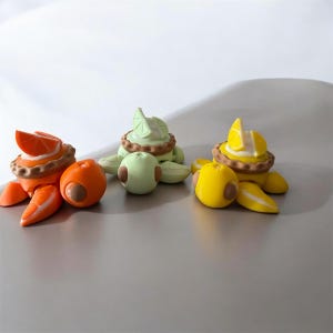 May include: Three whimsical fruit-themed turtle figurines. The turtles are orange, green, and yellow, each with a fruit slice on their backs. The orange turtle has an orange slice, the green turtle has a lime slice, and the yellow turtle has a lemon slice.