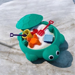 May include: A green turtle-shaped sandbox filled with white sand and colorful toys. The sandbox has a lid and features a variety of small plastic tools, including a rake, shovel, and bucket. The toys are in shades of red, yellow, orange, and blue.