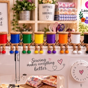 May include: A white sewing machine with the text Sewing makes everything better and a heart graphic. Eight wooden spools of thread in red, blue, green, yellow, purple, orange, white, and black are lined up on top, each with small articulated wooden legs.