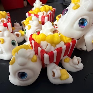 May include: A collection of whimsical, white popcorn-themed figurines. Each figure has a popcorn container with red and white stripes, filled with yellow and white popcorn. The figurines have colorful, detailed eyes and yellow accents.