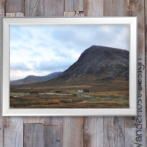 May include: A framed landscape print featuring a mountain range under a cloudy sky. The scene includes a small white building and a body of water in the valley. The print is set against a rustic wooden background.