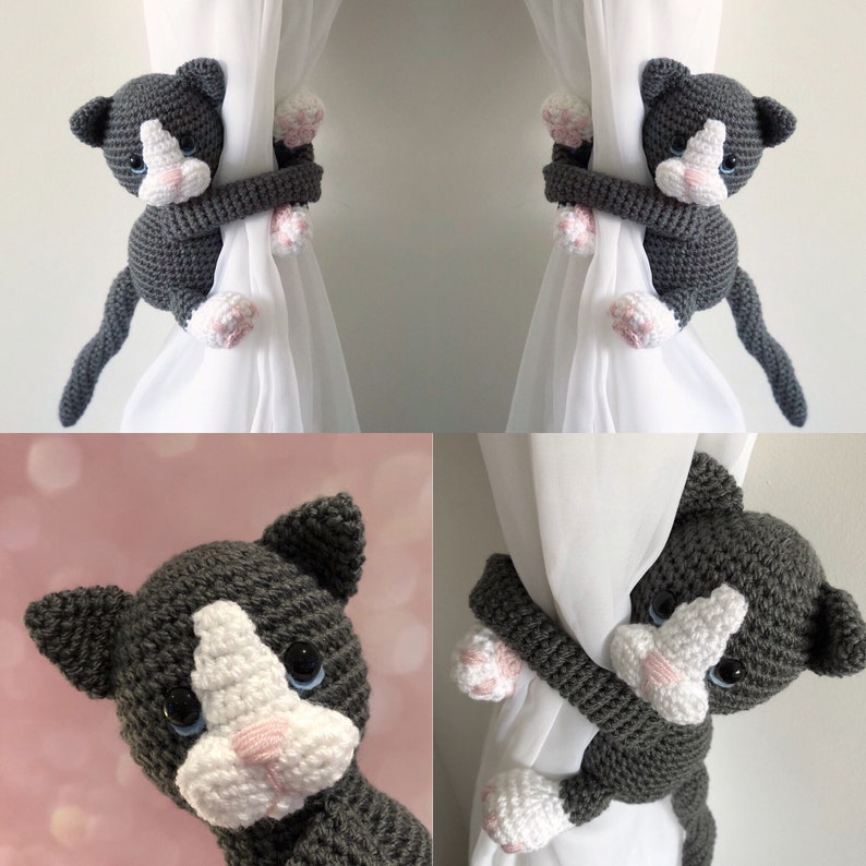 May include: Two gray and white crocheted cat curtain tiebacks. The cats are holding onto the curtain with their paws and have their tails hanging down.