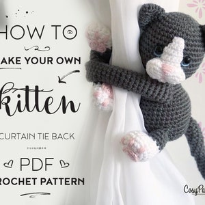May include: A crochet pattern for a gray and white kitten curtain tie back. The pattern is available as a PDF download. The image shows a finished kitten tie back holding a curtain.