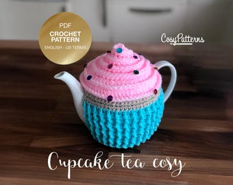 May include: A crocheted tea cosy shaped like a cupcake with pink icing and blue sprinkles. The cosy is on a white teapot. The text "Cupcake Tea Cozy" is written below the teapot.