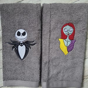 Pode incluir: Duas toalhas de m&atilde;o retangulares cinzentas com desenhos bordados. A toalha da esquerda mostra uma figura branca com cara de caveira num fato preto. A da direita apresenta uma personagem com cabelo vermelho, top amarelo e manga roxa.